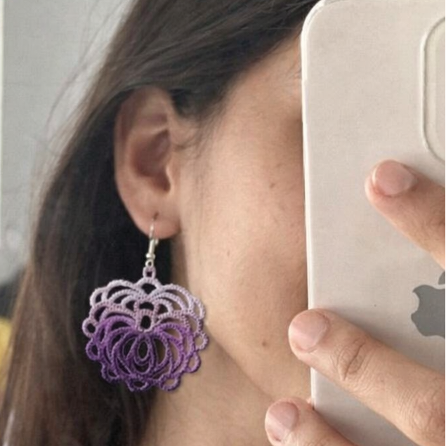 Person wearing a purple floral earring in a room with a bed and window.