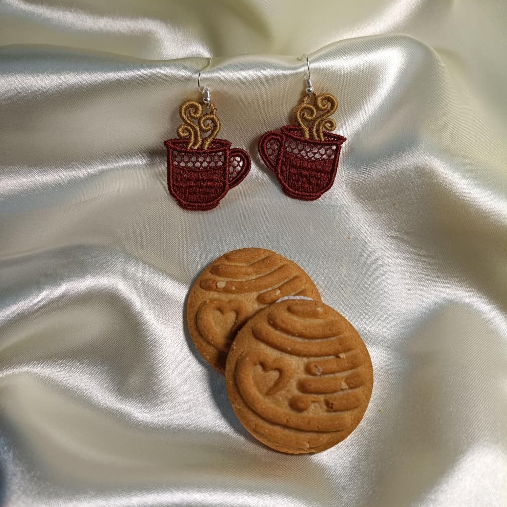 Earrings shaped like coffee cups and cookies on a satin surface