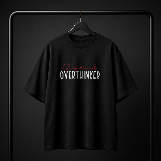 A black short-sleeve t-shirt hanging on a black hanger against a dark studio background. The shirt features a graphic text print on the chest reading 'Professional OVERTHINKER' in white and red lettering.