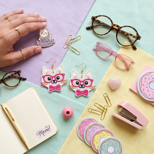 Cute cat embroidered earrings with pink glasses and bow placed on pastel desk setup with notebook, glasses, stationery, and aesthetic accessories, colorful flatlay scene