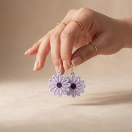 Hand holding a pair of purple flower earrings against a beige background
