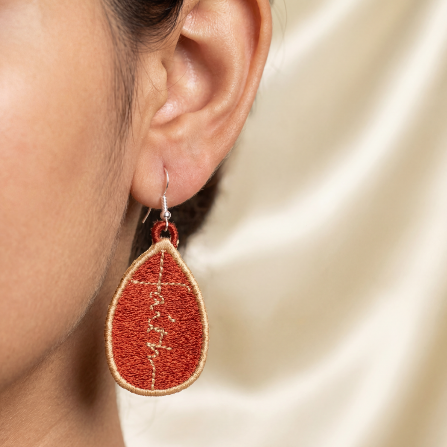 Teardrop-shaped earrings with red and gold design worn by a person on a beige background