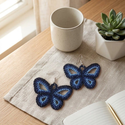 Blue butterfly earrings on a wooden table with a mug, plant, and notebook.