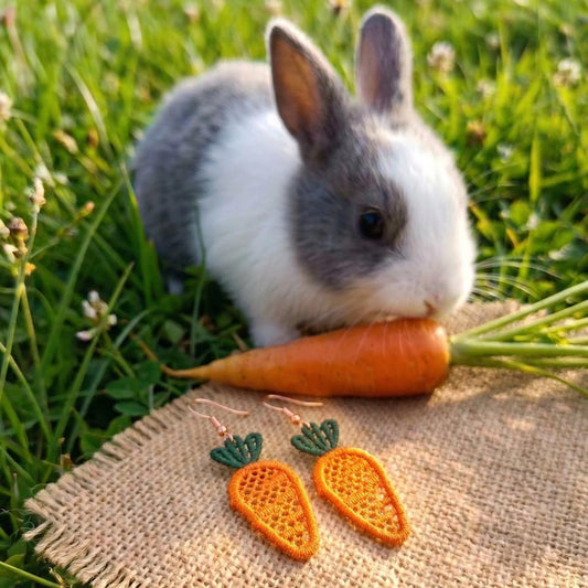 Small rabbit with a carrot on grass, next to carrot-shaped earrings on burlap fabric.